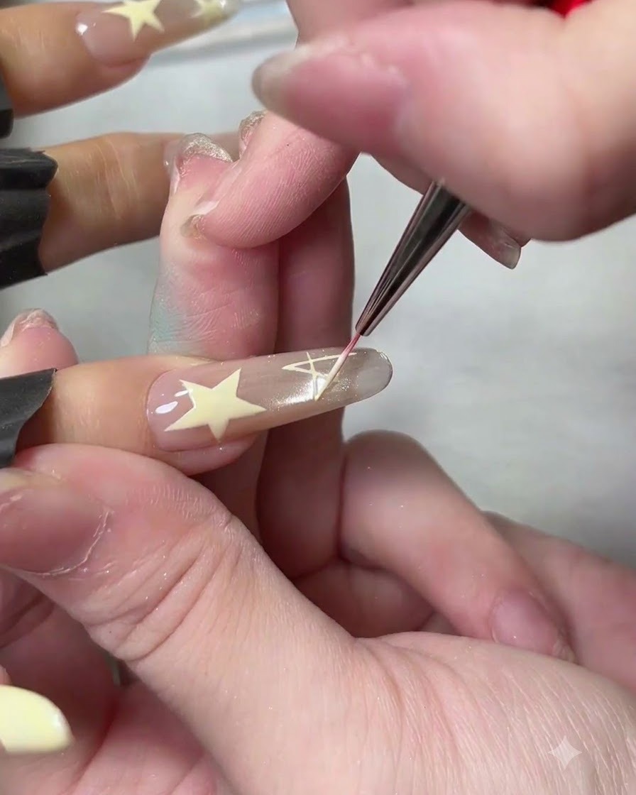 Un pinceau liner fin dessinant un design détoile pleine blanche sur un ongle artificiel de ton neutre.
