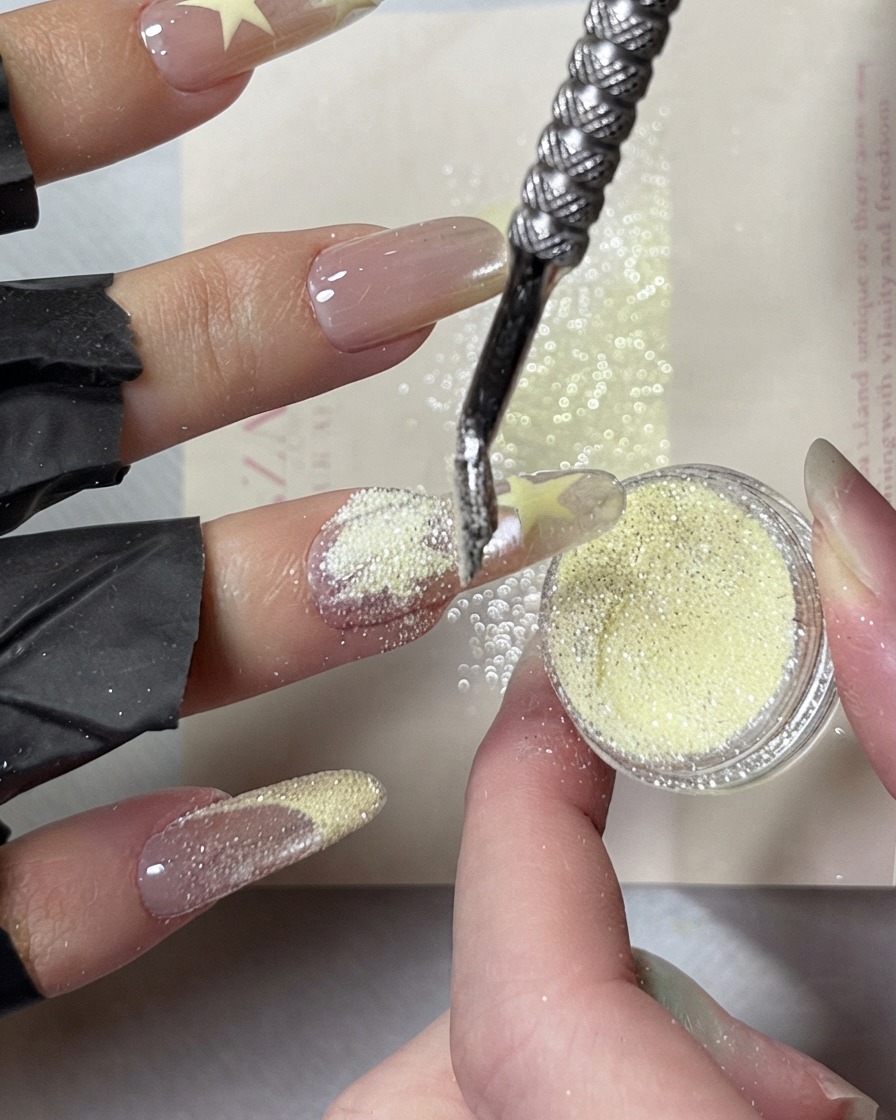 Un repousse-cuticules en métal ramassant et saupoudrant de la poudre de diamant concassée jaune crémeux brillante sur des motifs détoiles peints humides sur un ongle.