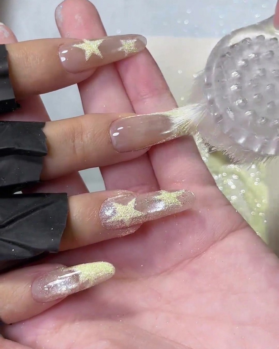Une brosse à épousseter ronde en plastique balayant la poudre jaune lâche des longs ongles nus en acrylique avec des détails détoiles texturées.