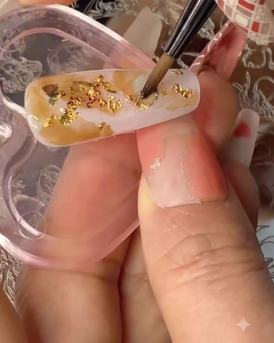 Pinceau de détail fin appliquant du gel solide transparent sur un ongle texturé avec de la feuille dor et des couleurs estompées.