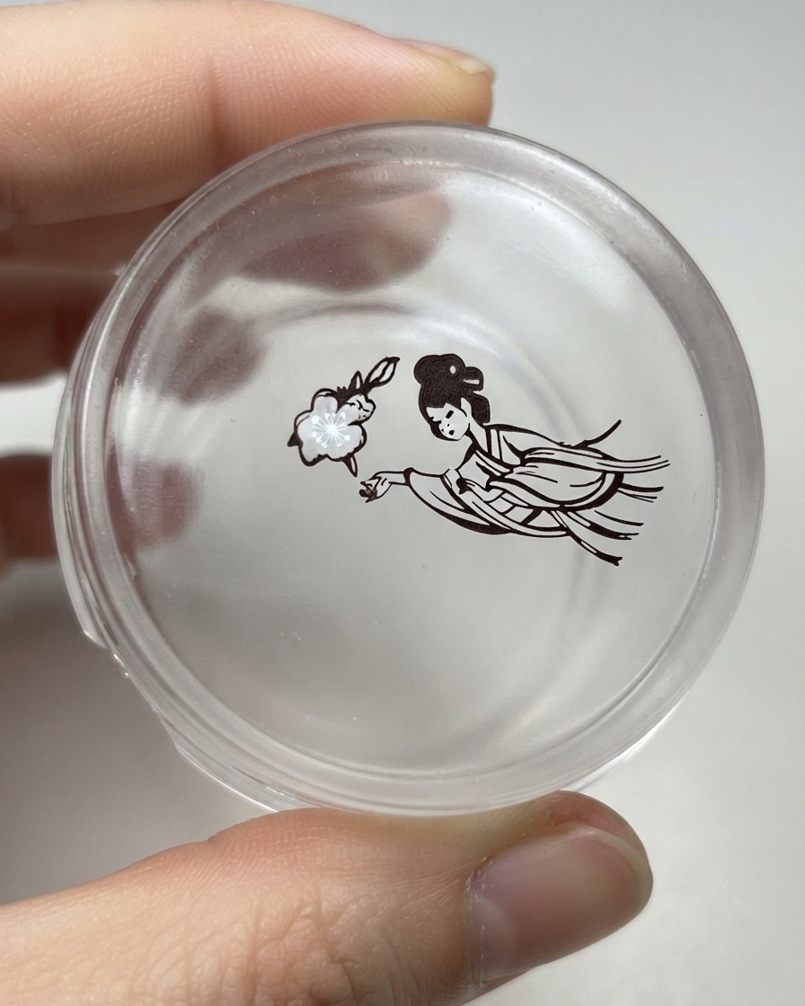 Une tête de tampon en silicone transparente tenant un design transféré dune fleur à côté dune figure traditionnelle féminine détaillée.
