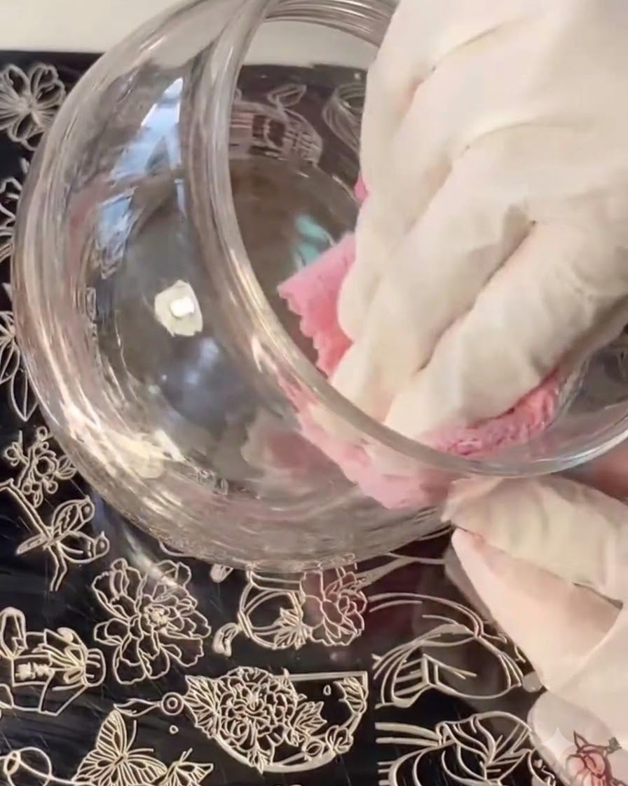 Une main gantée nettoyant ou essuyant un récipient en verre tandis que des plaques destampage aux motifs floraux se trouvent en dessous.