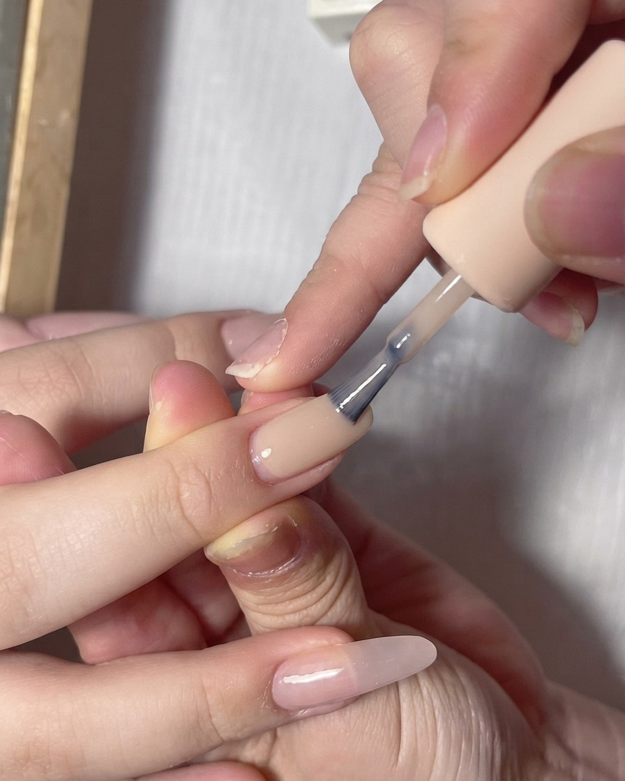 Un pinceau à vernis appliquant un gel de couleur plus claire et laiteux sur le bord libre dun ongle peint en nude rosé.