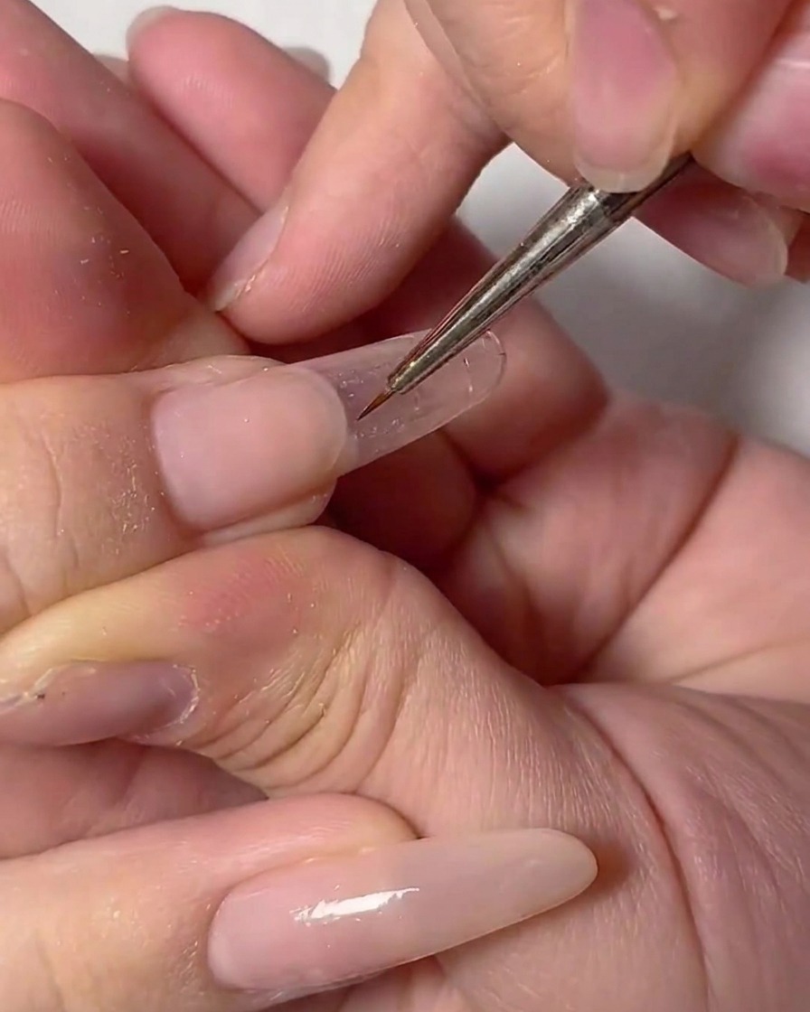 Gros plan dune main utilisant un pinceau fin de nail art pour dessiner un contour blanc délicat sur un ongle transparent.
