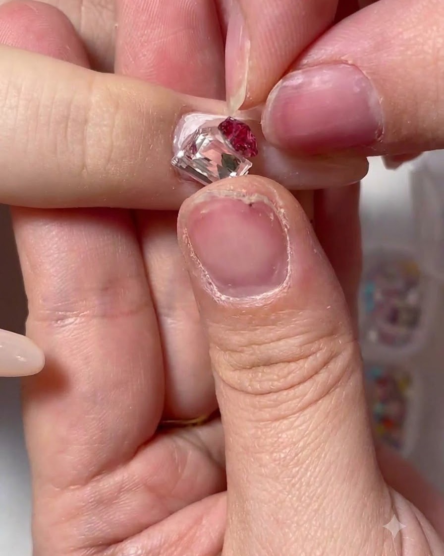 Une paire de brucelles posant un grand cristal carré et un groupe de strass floraux rouges sur un ongle décoré.