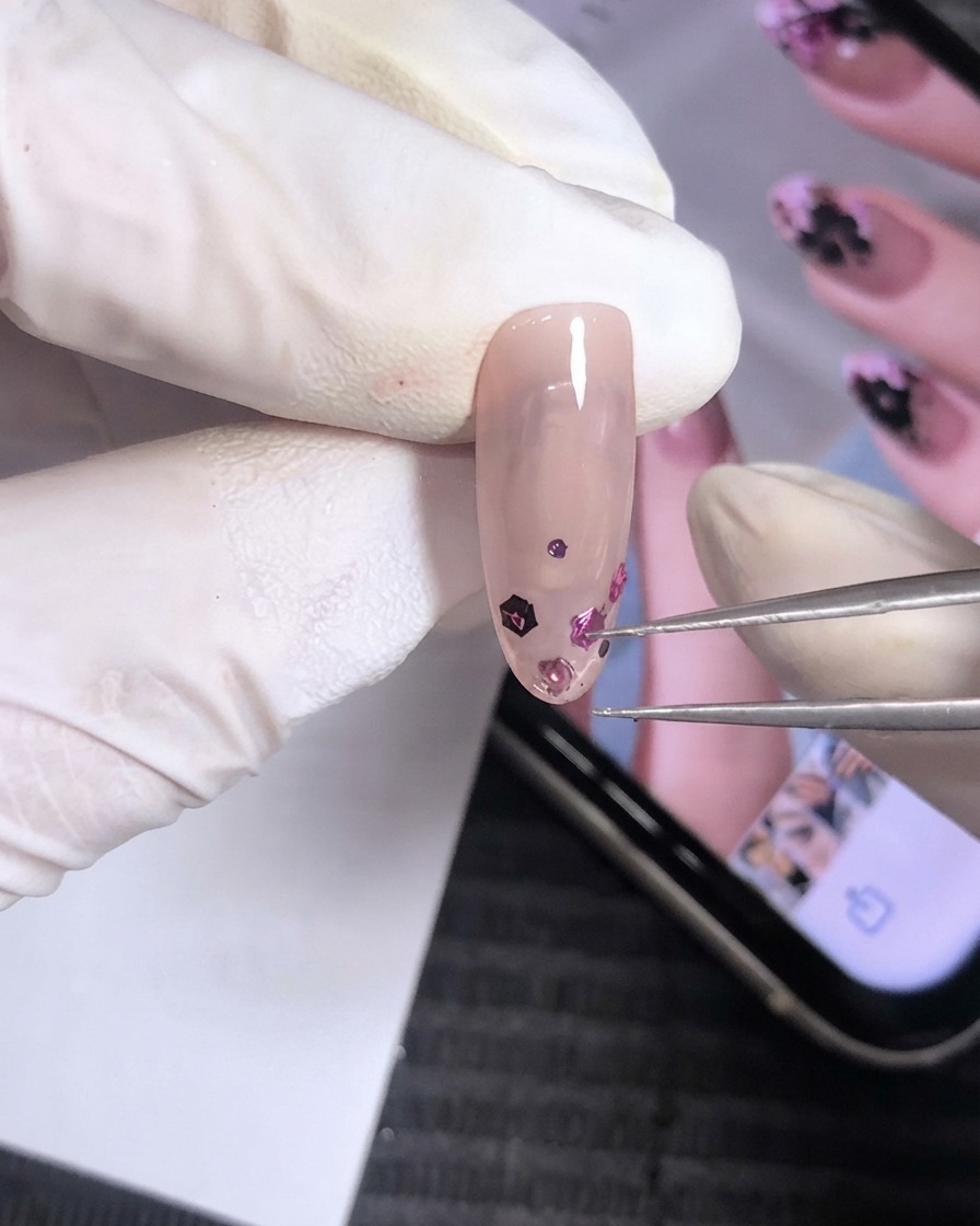 Une paire de fines pinces métalliques et un bâtonnet en buis ajustant des morceaux de sequins roses et violets sur un ongle en gel rose nude.