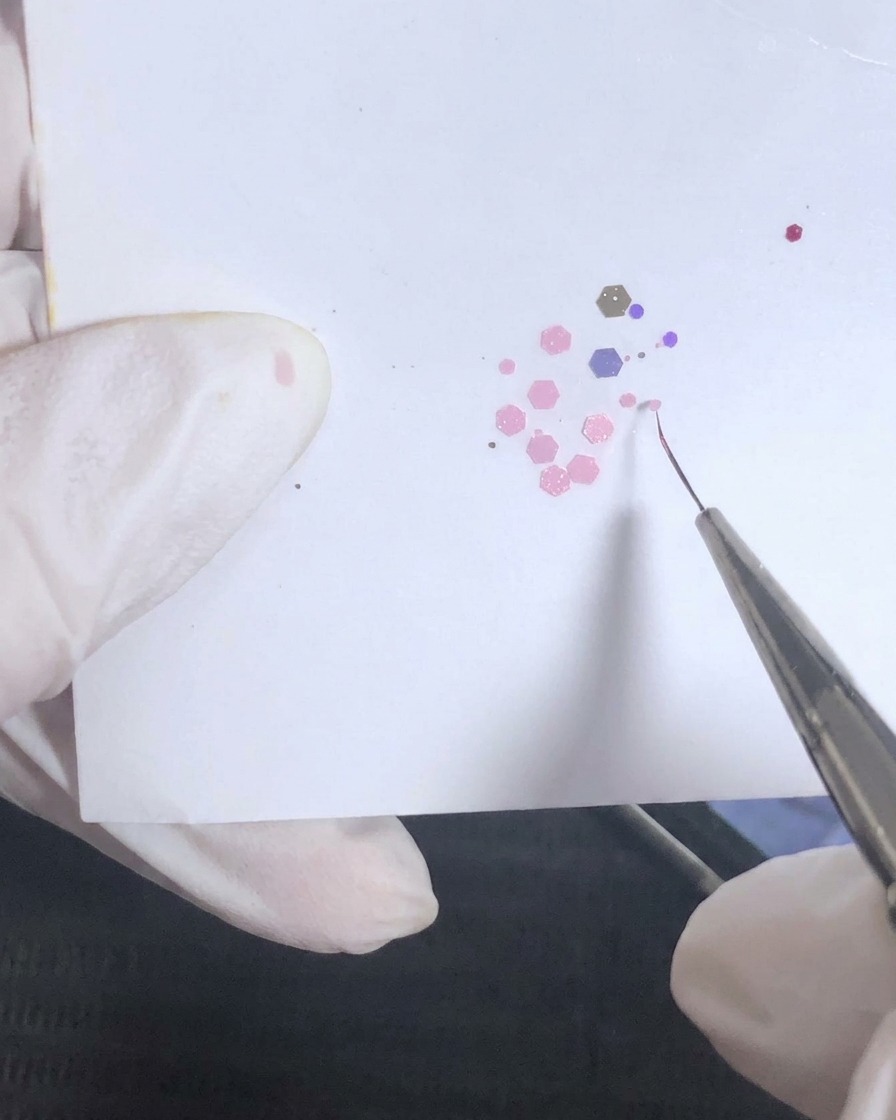 Un pinceau de détail fin ramassant de petits sequins hexagonaux roses sur une palette lors dun tutoriel de nail art.