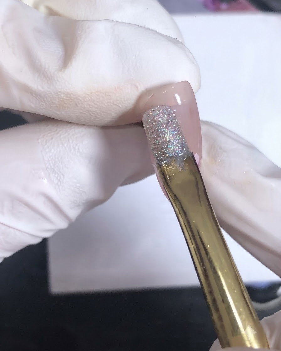 Un pinceau à manche doré appliquant un gel de scellement transparent sur un nail art texturé à sequins roses.