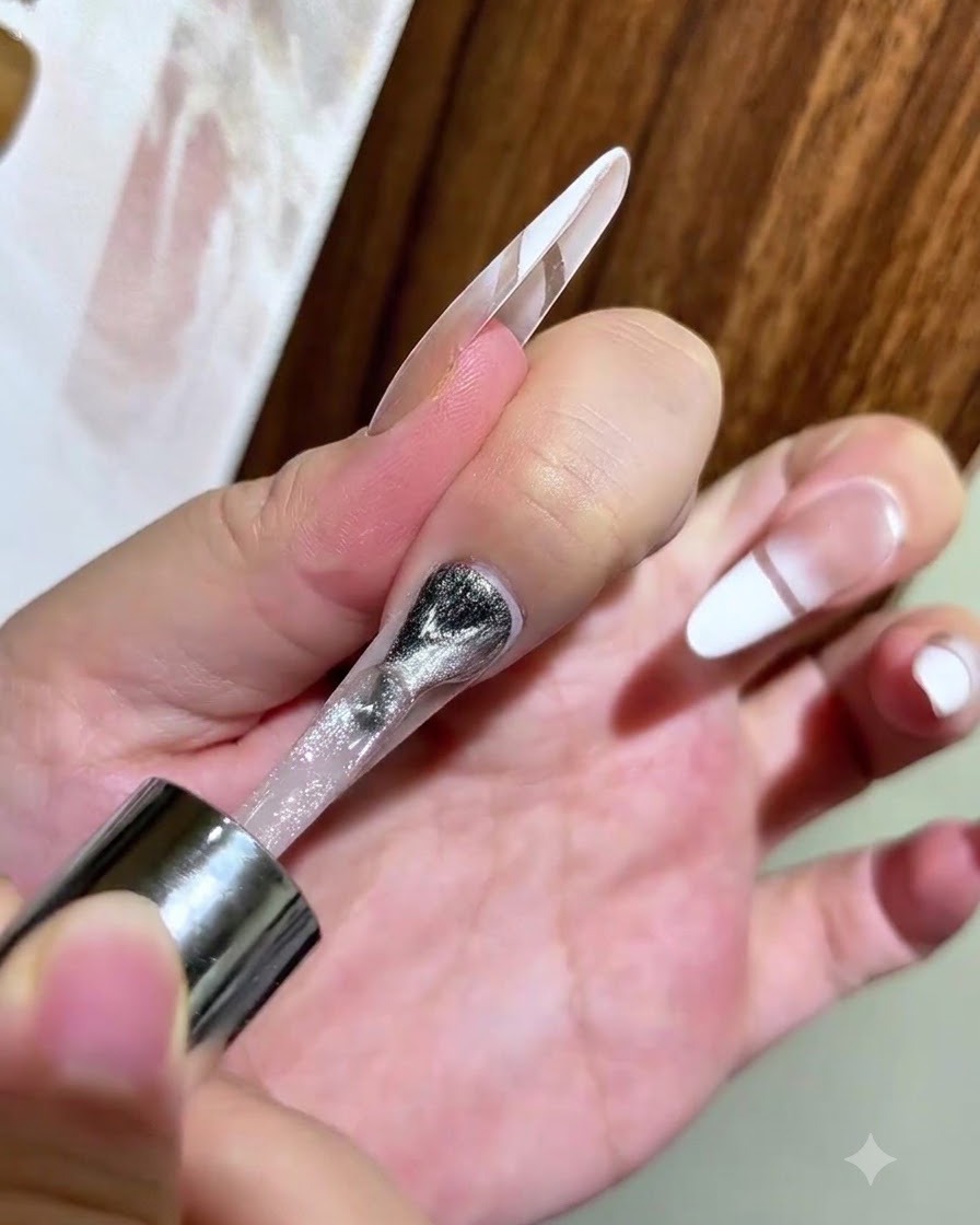 Un pinceau à vernis appliquant du gel magnétique cat eye argenté sur un ongle transparent avec un bout blanc dégradé.