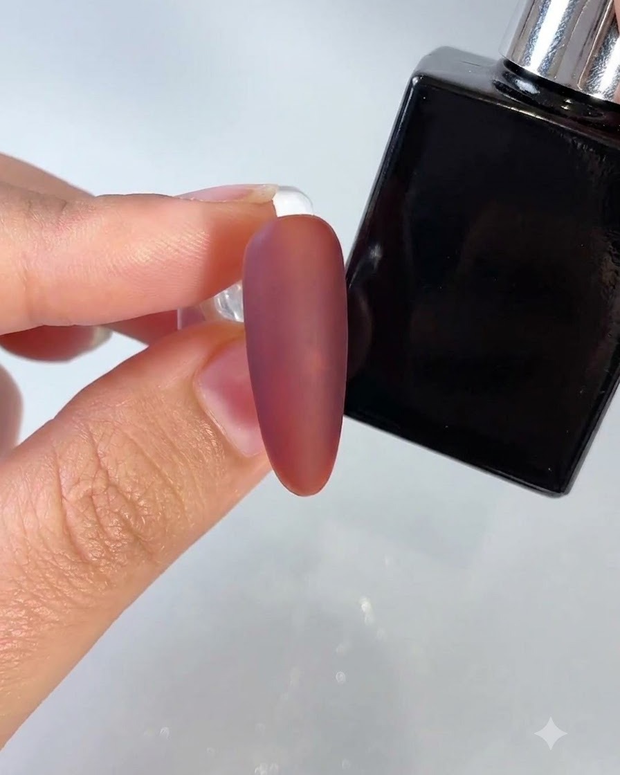 Un ongle rouge foncé à finition mate en cours dapplication avec un flacon de top coat spécialisé à proximité.