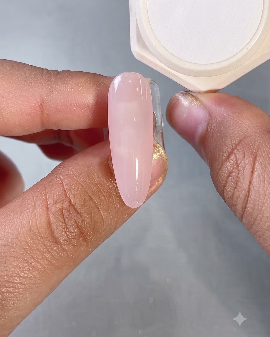 Un pinceau appliquant une couche de base gel rose laiteux translucide sur un ongle de forme ovale.