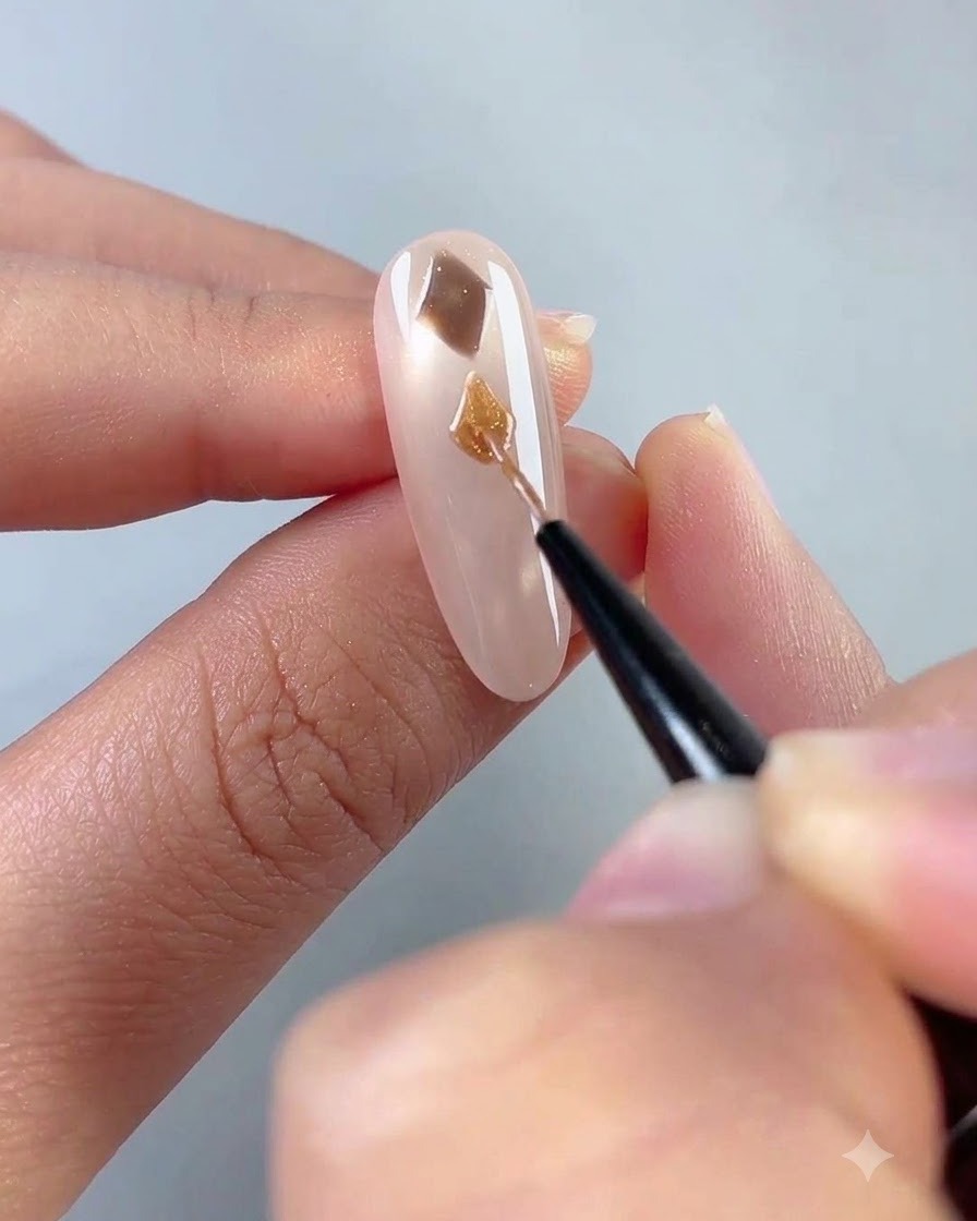 Gros plan dun pinceau de nail art fin peignant une forme de diamant marron sur un onglet rose diaphane.