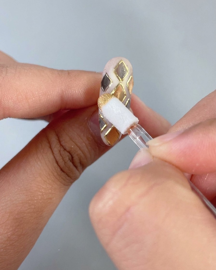 Une éponge de maquillage frottant de la poudre de chrome dorée sur un motif de grille en relief dun ongle peint.