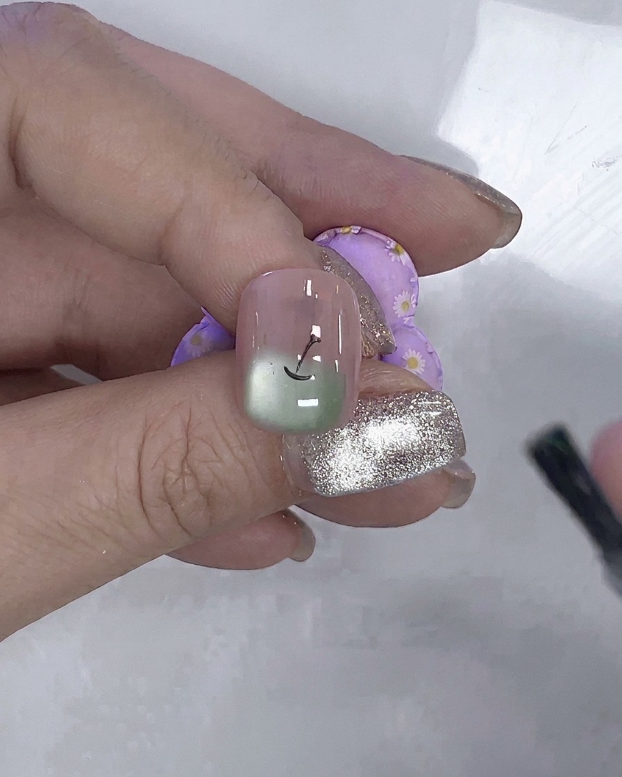 Un pinceau à ongle appliquant une couche de gel de renforcement transparent sur un ongle avec des détails de pomme peinte et cat-eye verte.